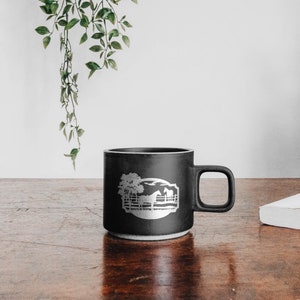 May include: A black ceramic mug with a white graphic of a horse, tree, and fence. The mug has a handle and a white base. The mug is sitting on a wooden surface.