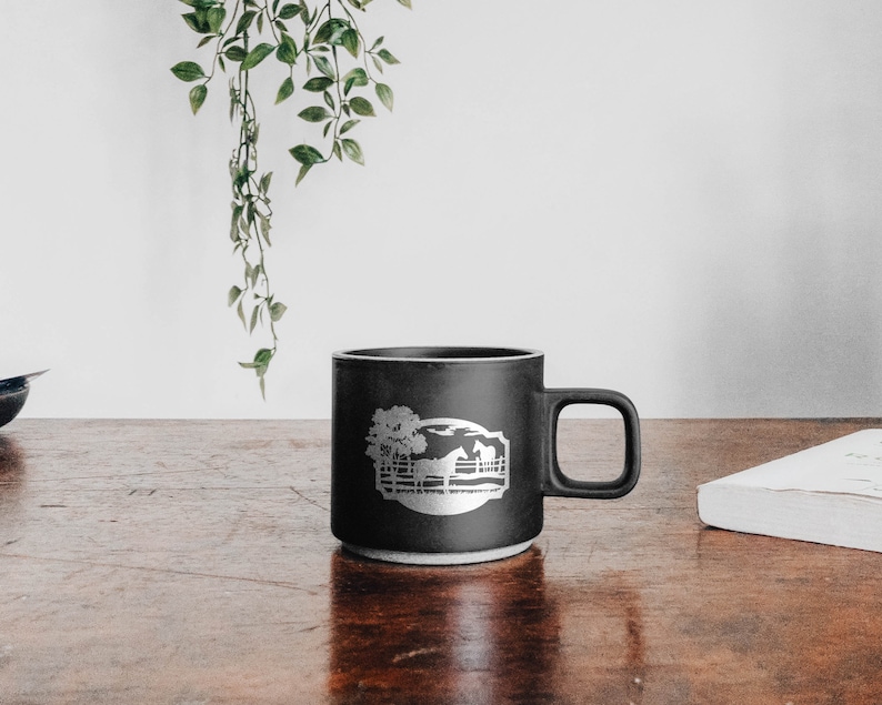 May include: A black ceramic mug with a white graphic of a horse, tree, and fence. The mug has a handle and a white base. The mug is sitting on a wooden surface.