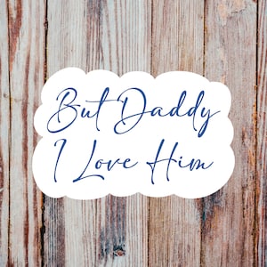 May include: White cloud-shaped sign with the text "But Daddy I Love Him" written in blue cursive font against a woodgrain background.