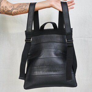 May include: Black backpack made from recycled materials. The bag features a rectangular shape with horizontal lines, adjustable black straps, and a small top handle. The straps are secured with silver-colored metal buckles. The bag is held up by a person.