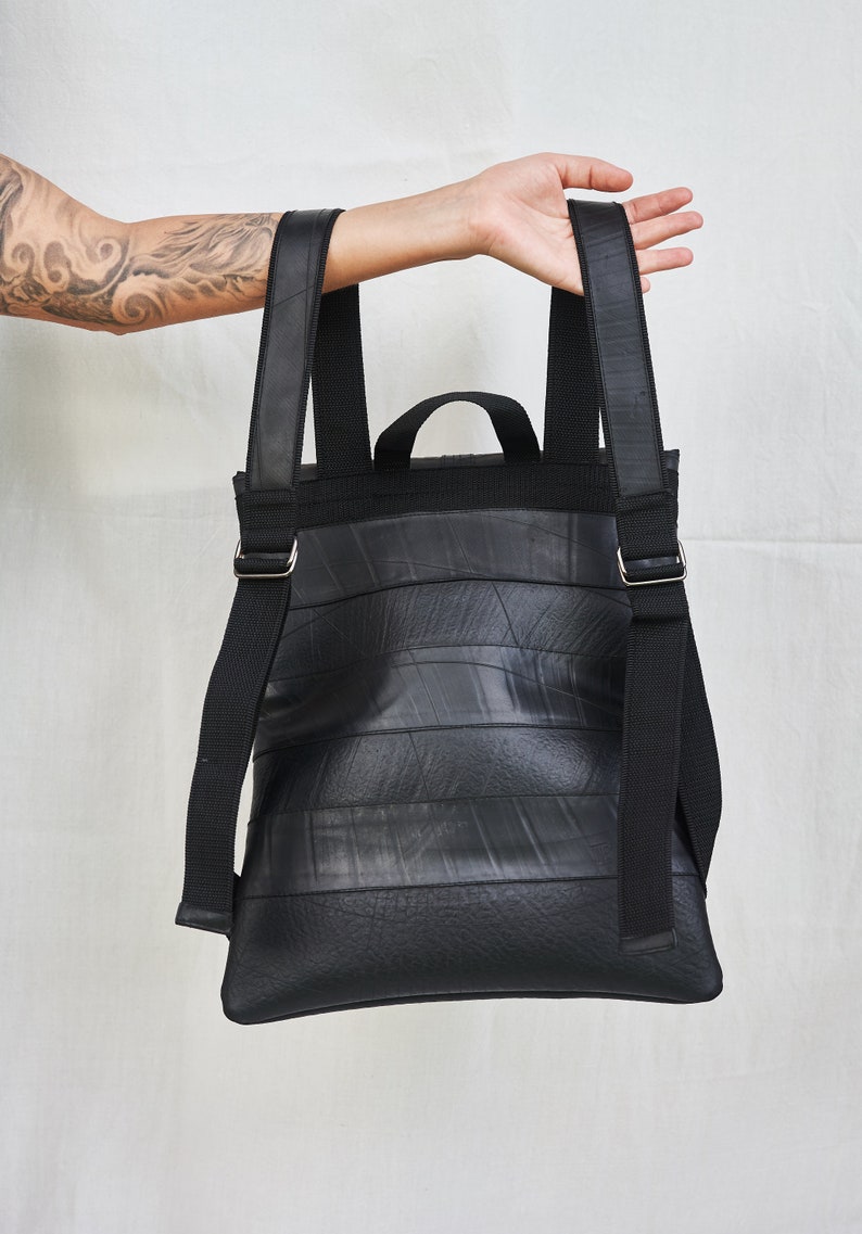 May include: Black backpack made from recycled materials. The bag features a rectangular shape with horizontal lines, adjustable black straps, and a small top handle. The straps are secured with silver-colored metal buckles. The bag is held up by a person.