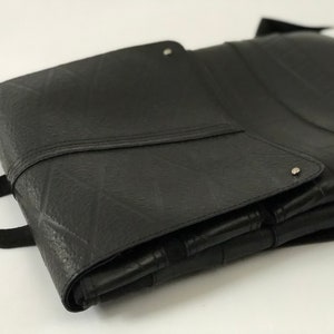 May include: Black, rectangular bag made from recycled tire material. The bag features a textured surface, a flap closure with a silver rivet, and a black strap handle. The bag is folded and appears to be empty.
