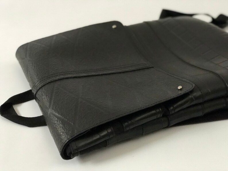 May include: Black, rectangular bag made from recycled tire material. The bag features a textured surface, a flap closure with a silver rivet, and a black strap handle. The bag is folded and appears to be empty.