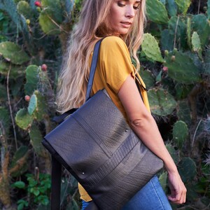 May include: Black rectangular backpack with a textured surface and a flap closure. The bag has a shoulder strap and is worn over the shoulder. The background features green plants and a person wearing a yellow shirt and blue jeans.