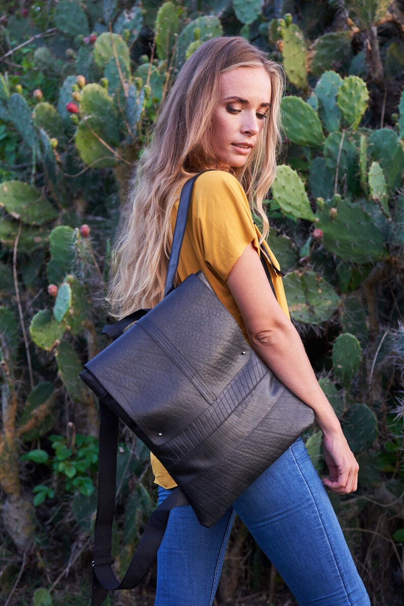 May include: Black rectangular backpack with a textured surface and a flap closure. The bag has a shoulder strap and is worn over the shoulder. The background features green plants and a person wearing a yellow shirt and blue jeans.