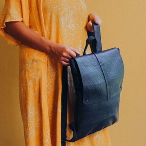 May include: A dark blue backpack with a flap closure and adjustable straps. The backpack is made of a textured material and has a rectangular shape. The bag is held by a person wearing a yellow dress.