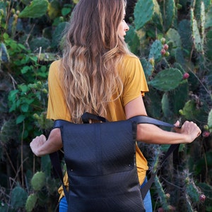 May include: Black backpack with adjustable straps. The backpack has a flap closure and a textured surface. The person in the image is wearing a yellow shirt and blue jeans. The background features green cacti.