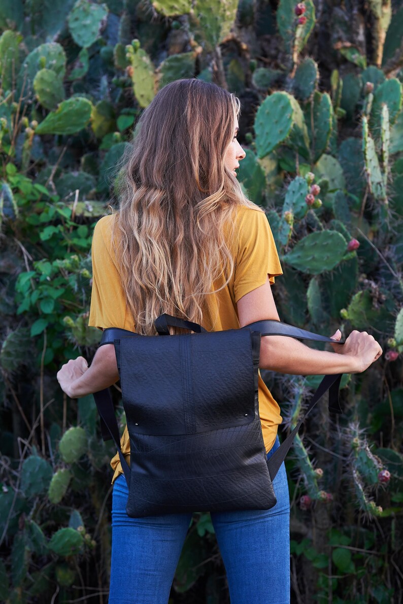 May include: Black backpack with adjustable straps. The backpack has a flap closure and a textured surface. The person in the image is wearing a yellow shirt and blue jeans. The background features green cacti.