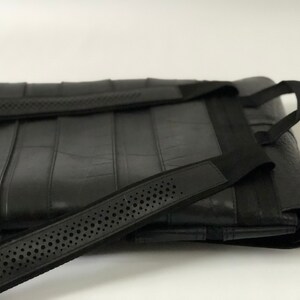 May include: Black tote bag with a textured surface and black straps. The bag has a rectangular shape with vertical seams and a strap with small, evenly spaced holes. The straps are attached to the bag with black fabric.