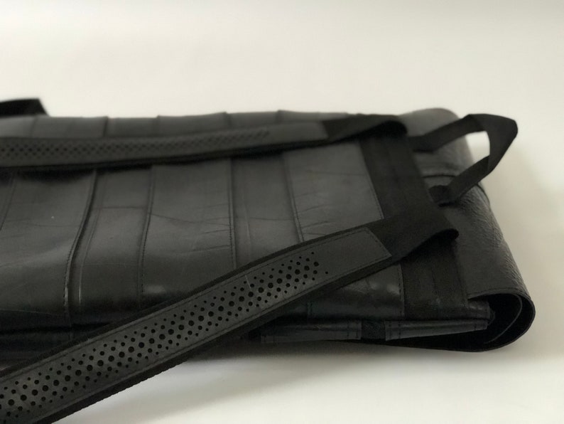 May include: Black tote bag with a textured surface and black straps. The bag has a rectangular shape with vertical seams and a strap with small, evenly spaced holes. The straps are attached to the bag with black fabric.