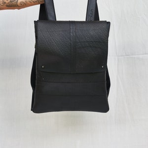 May include: Black backpack with a textured surface, held up against a white background. The bag has a flap closure and adjustable straps. The straps are black and the bag appears to be made of a durable material.