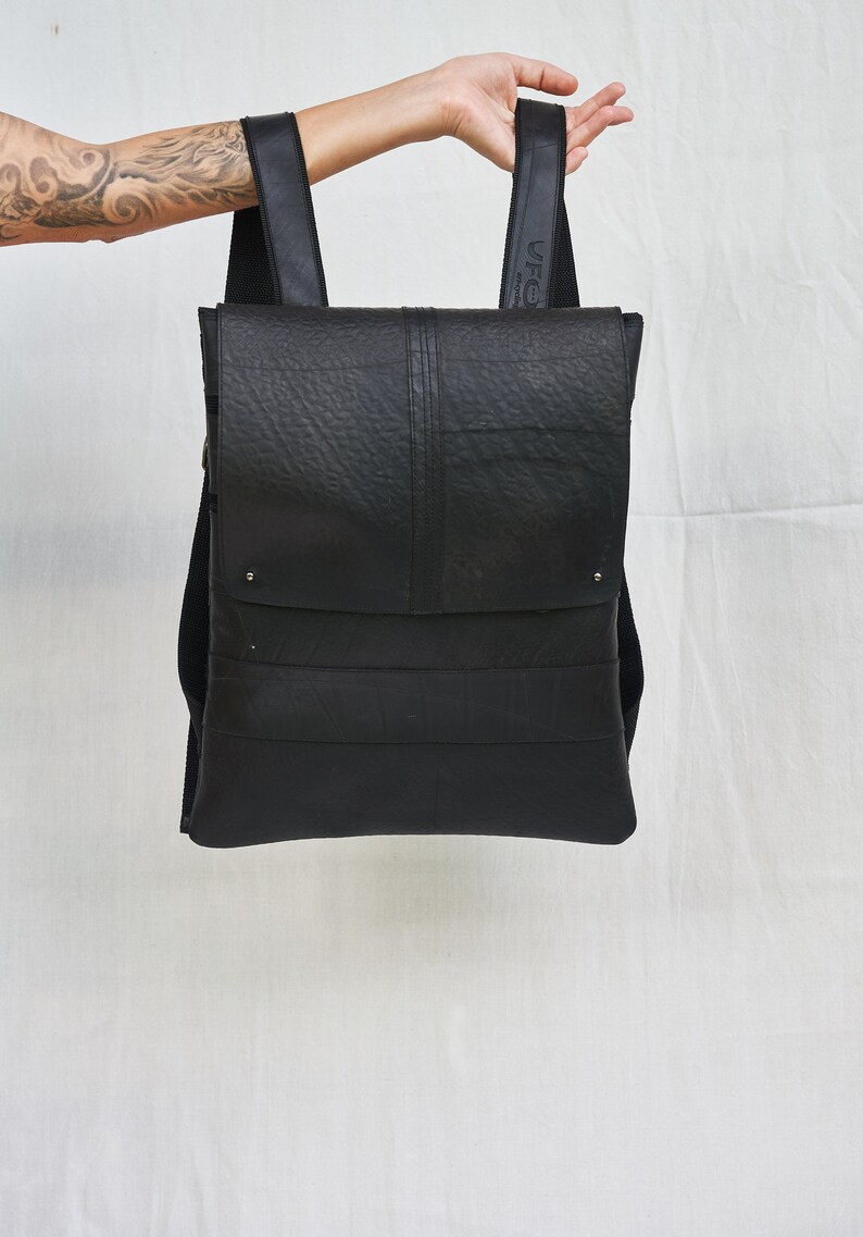 May include: Black backpack with a textured surface, held up against a white background. The bag has a flap closure and adjustable straps. The straps are black and the bag appears to be made of a durable material.