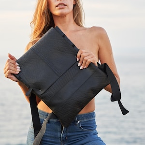 May include: Black rectangular messenger bag with a flap closure and adjustable shoulder strap. The bag has a textured surface and is held against a background of water and sky. The bag is made of a durable material.