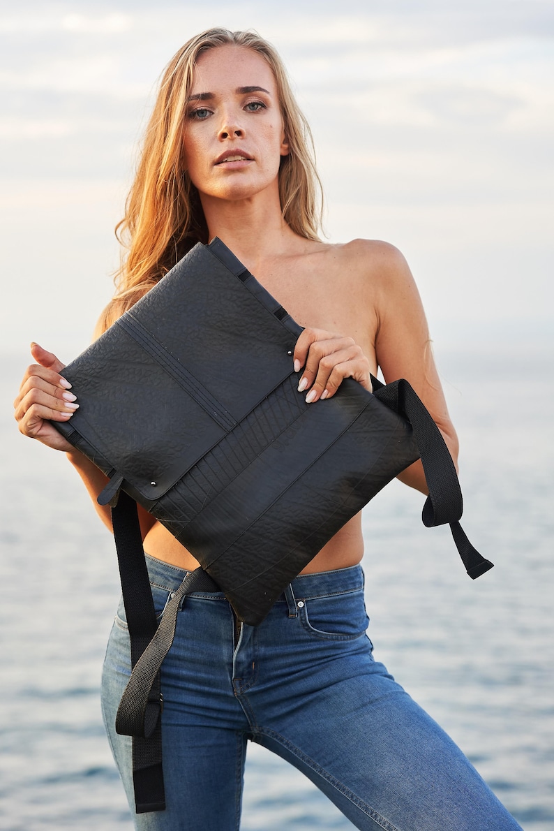 May include: Black rectangular messenger bag with a flap closure and adjustable shoulder strap. The bag has a textured surface and is held against a background of water and sky. The bag is made of a durable material.