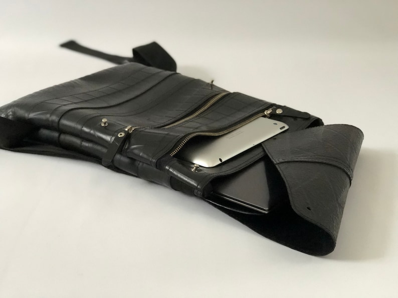 May include: Black messenger bag made from recycled tire material. The bag features a zippered main compartment, a front pocket, and a shoulder strap. A tablet and laptop are visible inside the bag.