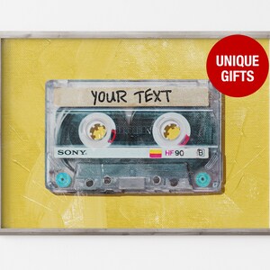 以下が含まれることがあります： 黄色の背景に、ビンテージのカセットテープ。テープには「YOUR TEXT」と書かれています。テープは銀色のフレームに入っています。