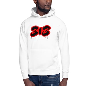 Puede incluir: Sudadera con capucha blanca con bolsillo delantero y cordón. La sudadera presenta el número "313" en negro con contorno rojo y efecto de pintura negra goteando.
