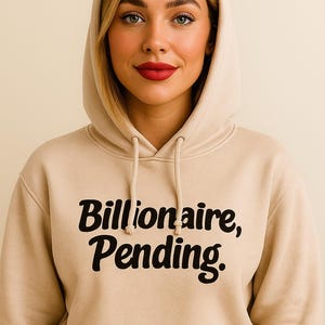 Puede incluir: Una sudadera con capucha beige con el texto "Billionaire, Pending" en negro. La sudadera con capucha tiene una capucha con cordón y un bolsillo delantero. La persona que lleva la sudadera con capucha tiene el pelo rubio y lápiz labial rojo.