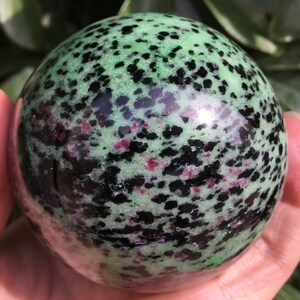 Puede incluir: Una esfera de piedra preciosa verde, negra y rosa. La esfera está pulida y tiene un patrón moteado.