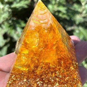 Pode incluir: Uma pirâmide de cristal amarelo transparente com manchas douradas e um padrão em espiral no seu interior. A pirâmide é feita de citrino e é uma escolha popular para meditação e cura energética.
