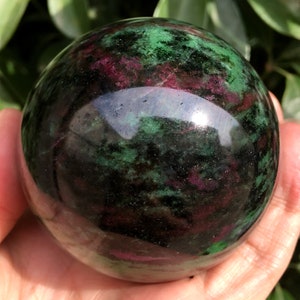 May include: A polished green and black sphere with pink and red inclusions. The sphere is made of a natural stone and has a smooth, glossy finish.