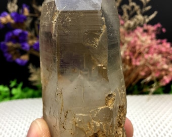 Puede incluir: Un gran cristal de cuarzo transparente con una base natural rugosa. El cristal es puntiagudo en la parte superior y tiene una apariencia translúcida y vidriosa.