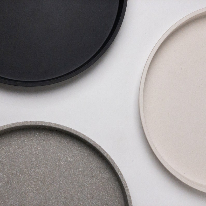 May include: Three round trays in different colours and textures. The tray on the left is black, the tray on the right is white, and the tray in the centre is grey with a speckled texture.