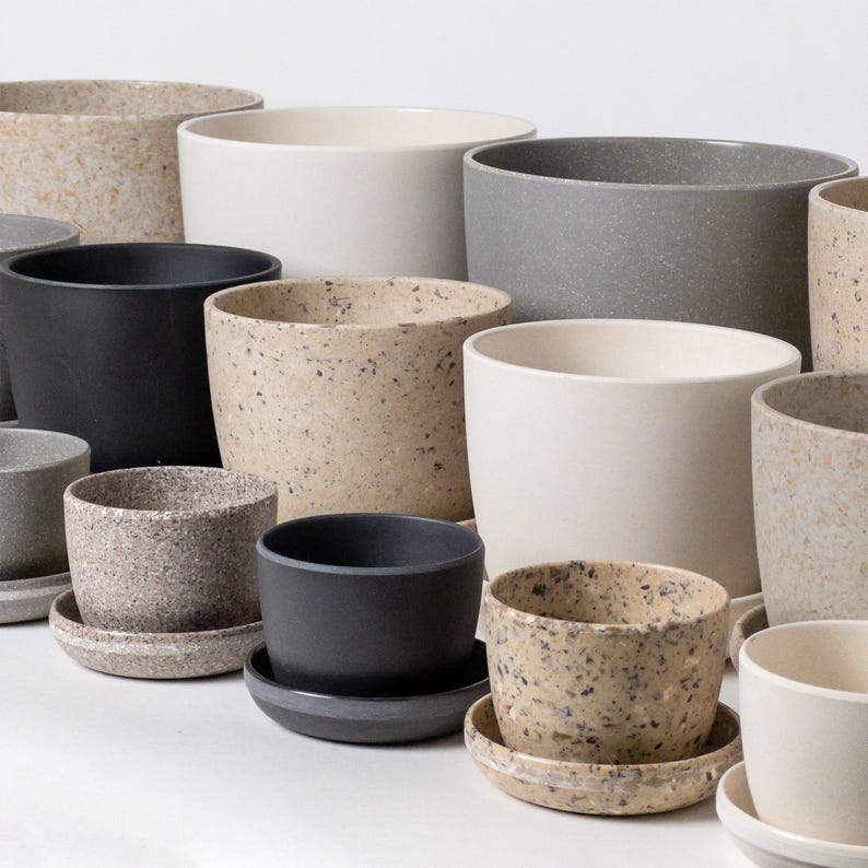 Op de afbeelding: Een verzameling gespikkelde keramische potten in verschillende maten en kleuren, waaronder beige, grijs en zwart. De potten zijn in een verspringende opstelling geplaatst, met de grootste pot achterin en de kleinste pot vooraan.