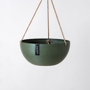 Op de afbeelding: Een donkergroene, komvormige hangende plantenbak met een gladde, matte afwerking. De plantenbak hangt aan drie dunne, lichtbruine touwen die samenkomen in een centraal punt. Een klein zwart label bevat de witte tekst KANSO.
