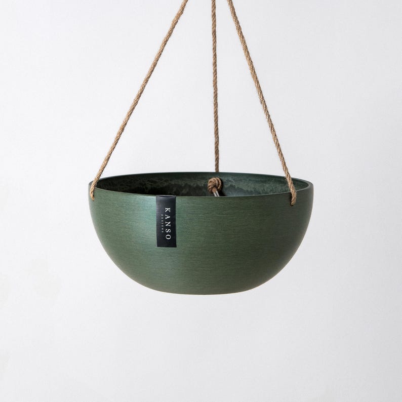 Op de afbeelding: Een donkergroene, komvormige hangende plantenbak met een gladde, matte afwerking. De plantenbak hangt aan drie dunne, lichtbruine touwen die samenkomen in een centraal punt. Een klein zwart label bevat de witte tekst KANSO.