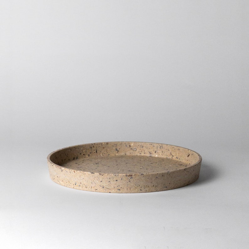 May include: A round, speckled, beige tray made of concrete. The tray has a slightly raised rim.