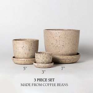 Op de afbeelding: Een set van drie gespikkelde beige keramische potten met bijpassende schotels. De potten hebben een diameter van 7,6 cm, 12,7 cm en 17,8 cm. "3 Piece Set Made From Coffee Beans" is afgedrukt onder de potten.