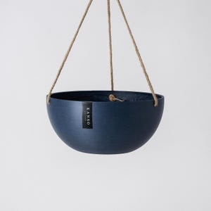 Op de afbeelding: Een donkerblauwe, komvormige hangende plantenbak met een subtiele horizontale geborstelde textuur. De plantenbak hangt aan drie beige touwen van jute en is voorzien van een verticaal zwart rechthoekig label met de witte tekst KANSO DESIGNS.