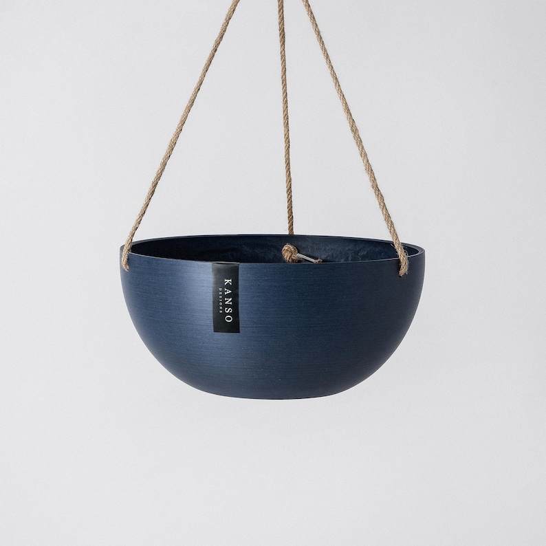 Op de afbeelding: Een donkerblauwe, komvormige hangende plantenbak met een subtiele horizontale geborstelde textuur. De plantenbak hangt aan drie beige touwen van jute en is voorzien van een verticaal zwart rechthoekig label met de witte tekst KANSO DESIGNS.