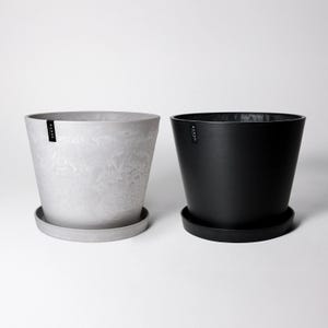 May include: Two plant pots, one grey and one black, with a textured finish. Both pots have a saucer and the word "KANZA" printed on the side.