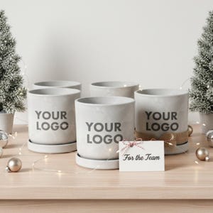 May include: Five light grey cylindrical planters with saucers, each bearing the text "YOUR LOGO". A small card reading "For the Team" is tied with twine. The scene is decorated with small Christmas trees and baubles, creating a festive display.