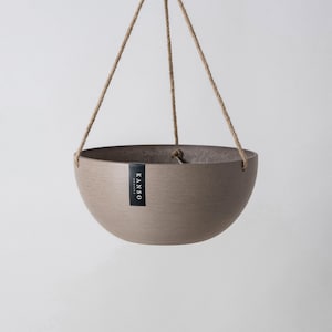 Op de afbeelding: Een komvormige hangende plantenbak in een gedempte taupekleur met een getextureerde afwerking. De plantenbak hangt aan drie dunne, lichtbruine touwen die aan de rand zijn bevestigd. Een klein zwart rechthoekig label met de witte tekst KANSO DESIGNS is bevestigd.