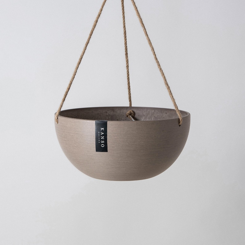 Op de afbeelding: Een komvormige hangende plantenbak in een gedempte taupekleur met een getextureerde afwerking. De plantenbak hangt aan drie dunne, lichtbruine touwen die aan de rand zijn bevestigd. Een klein zwart rechthoekig label met de witte tekst KANSO DESIGNS is bevestigd.