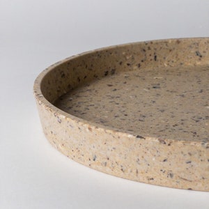 May include: A round, beige tray made of speckled terrazzo. The tray has a slightly raised rim and a smooth, polished surface.