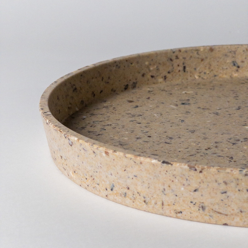 May include: A round, beige tray made of speckled terrazzo. The tray has a slightly raised rim and a smooth, polished surface.