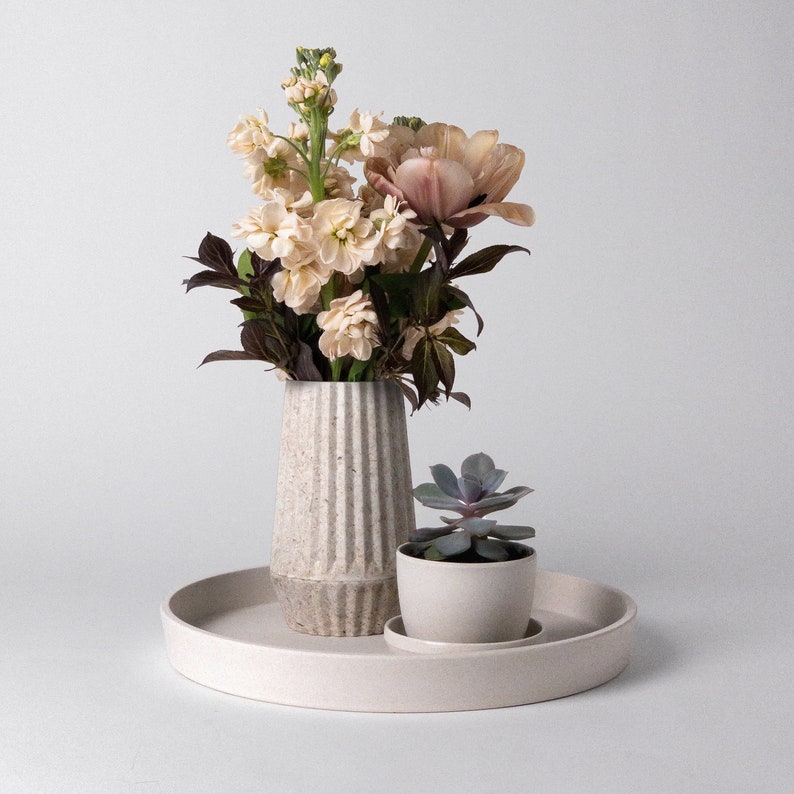 May include: A white ceramic vase with a textured surface holds a bouquet of light pink and cream flowers. A small succulent plant in a white ceramic pot sits on a white circular tray.
