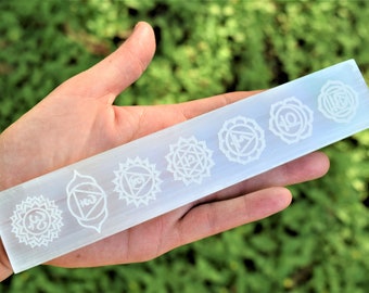 May include: White selenite wand with seven chakra symbols engraved on the surface. The symbols are in a row, starting with the root chakra and ending with the crown chakra.