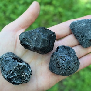 May include: Four black obsidian stones of varying sizes and shapes, with a smooth, polished surface. The stones are displayed on a hand, with a green background.