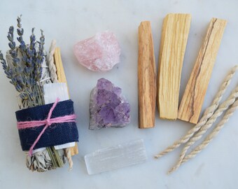 May include: A smudging kit with white sage, lavender, palo santo sticks, selenite, amethyst, and rose quartz crystals. The kit is tied with pink twine and wrapped in blue fabric.