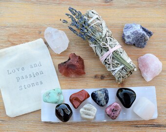 May include: A white cloth bag with the text "Love and passion stones" on it. The bag is surrounded by various crystals and stones, including clear quartz, rose quartz, amethyst, red jasper, and a bundle of sage.