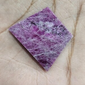 Könnte beinhalten: Ein rautenförmiger, polierter Edelstein in leuchtendem Rosa, durchzogen von weißen und schwarzen Einschlüssen. Die Oberfläche des Steins weist eine strukturierte Erscheinung auf, die natürliche Muster zeigt. Dieser Edelstein wird wahrscheinlich für Schmuck oder Dekorationszwecke verwendet.