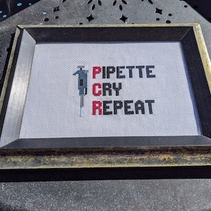 May include: A framed cross-stitch with a black and white illustration of a person holding a pipette. The text reads "Pipette Cry Repeat" in black and red lettering.