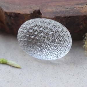 May include: A clear glass cabochon with a textured surface. The cabochon has a round shape and is approximately 2 inches in diameter.