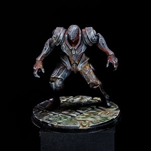 Könnte beinhalten: Eine detaillierte Miniaturfigur einer humanoiden Kreatur, die eine silberne und braune Rüstung trägt. Die Figur ist in einer Kampfhaltung mit erhobenen Armen und geballten Fäusten dargestellt. Die Figur steht auf einem runden Sockel mit einer strukturierten Oberfläche.