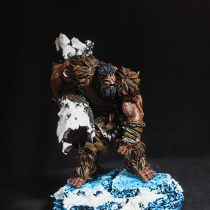 Puede incluir: Figura en miniatura detallada de una criatura humanoide grande y musculosa con pelaje marrón y barba. La figura se encuentra sobre una base nevada azul y blanca, con nieve en los hombros y los brazos. La figura está pintada con detalles intrincados.